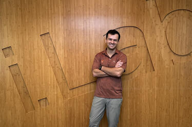 Andrew Schoka smiling with arms crossed, standing in front of a wooden wall with “i.Lab” engraved in large letters.