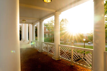 Sunlight streams through white columns on a brick veranda, with a view of a tree-lined campus and red-brick buildings in the background.